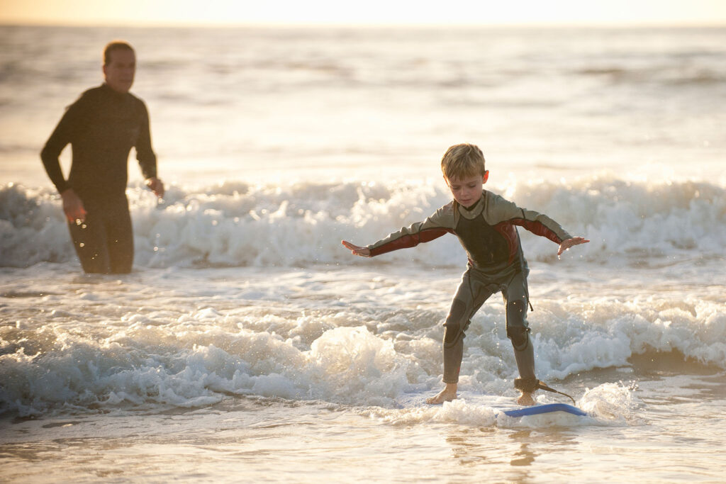 A boy in a wetsuit surfs a small wave as Dad looks on.