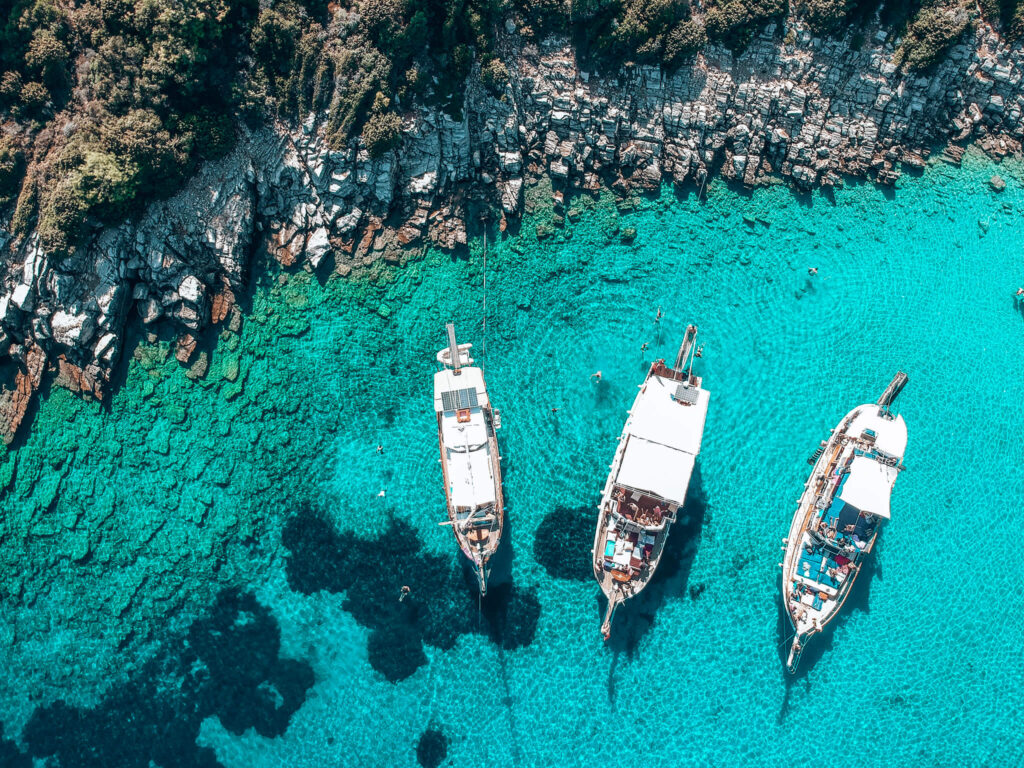 Three charter boats sit anchored next to shore while customers swim in the crystal-clear water of Orak Island.