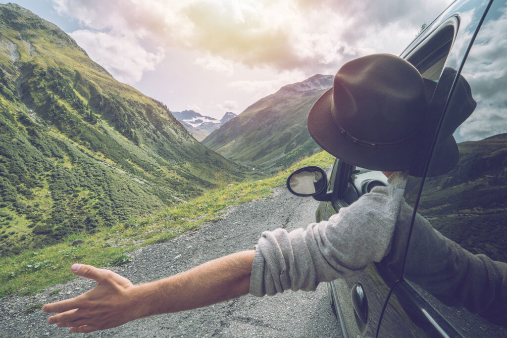 A man spreads his arms from a car window while admiring green mountains.