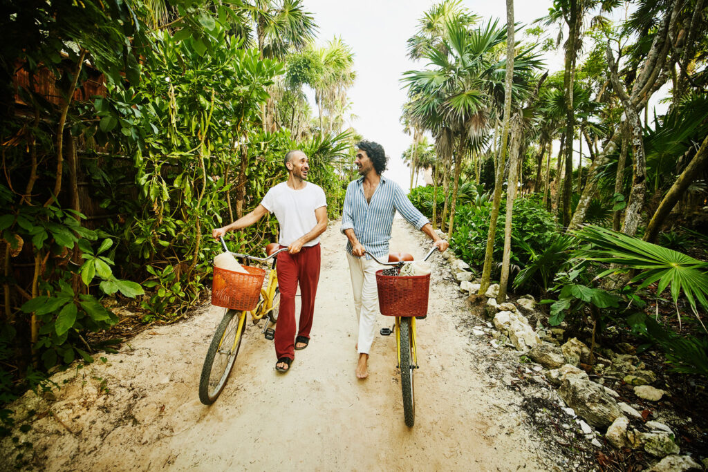 Two men walk their bikes up the path from a tropical beach.