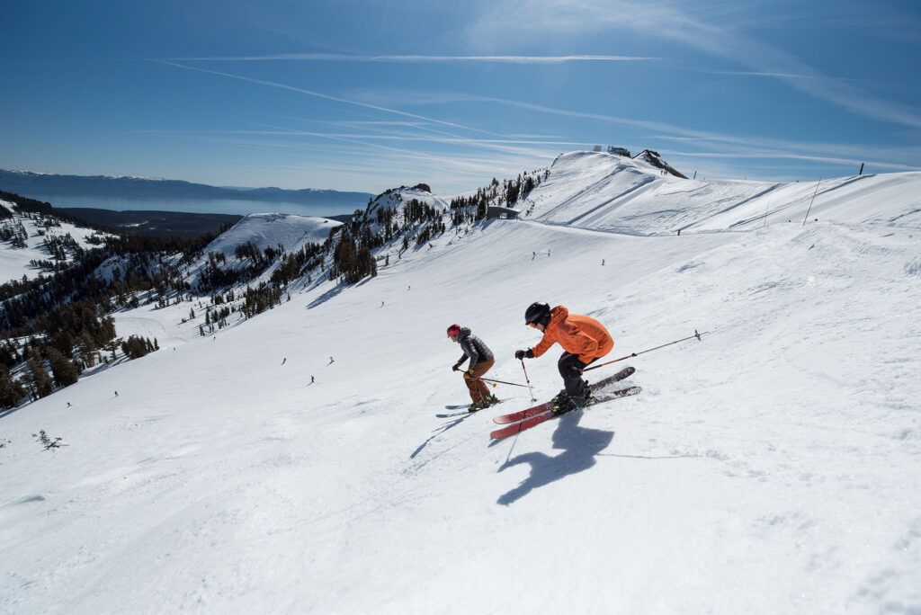 Two snow skiers zoom down broad mountain slopes in Lake Tahoe.