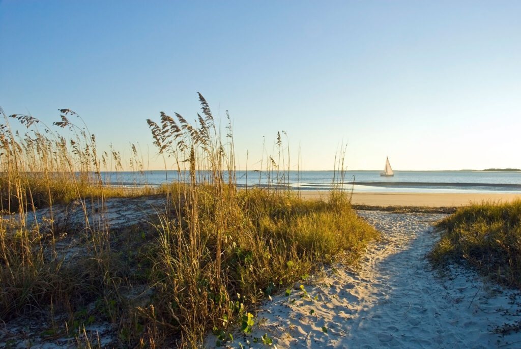 A sandy path through the sea oats and dunes leads to the beach, where a sailboat eases by.