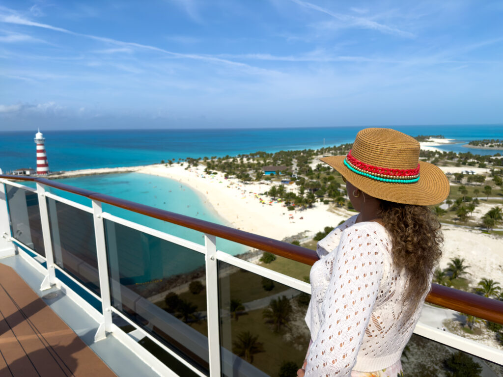 A beautiful woman stands on the railing of a cruise ship, taking in the stunning views of the clear blue sea on a bright, sunny day. The vibrant waters stretch out to the horizon, while a picturesque lighthouse stands on the beach, completing this tranquil Bahamian scene. This image captures the essence of relaxation, adventure, and the beauty of tropical escapes. Perfect for showcasing luxury travel, serene landscapes, and summer vacations